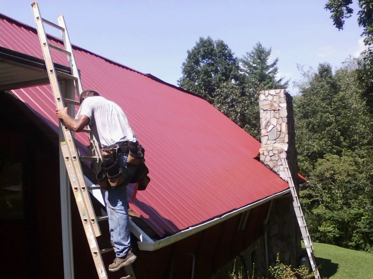 Roofer on a ladder beside a red metal roof and stone chimney — Soffit & Fascia Repair by Roofing Brothers in Wynnewood